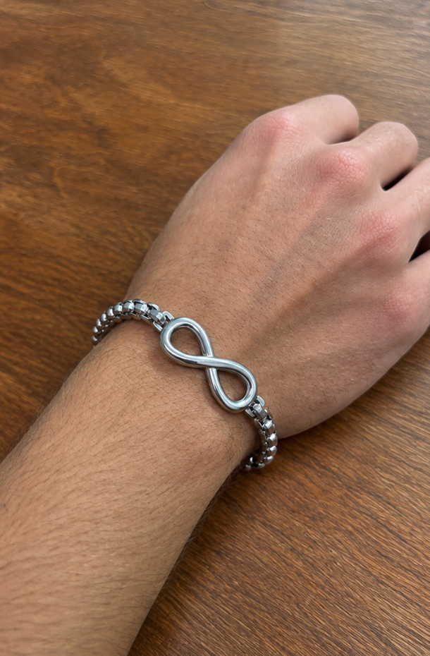 Silver infinity bracelet with box chain on a man's wrist, resting on a wooden table